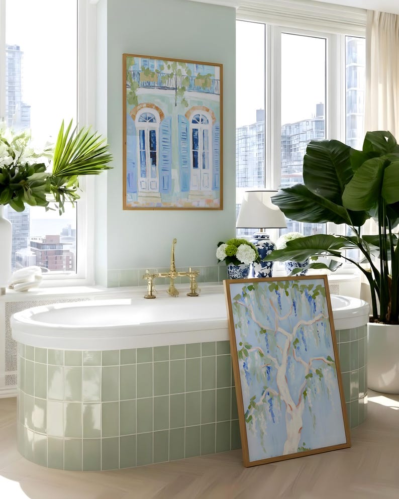 Puede incluir: Un ba&ntilde;o con una ba&ntilde;era ovalada blanca y grifos de lat&oacute;n, rodeado de azulejos verde claro. Dos cuadros enmarcados muestran escenas arquitect&oacute;nicas y bot&aacute;nicas. Tambi&eacute;n se ven una gran planta en maceta y una l&aacute;mpara blanca con una base estampada en azul y blanco.