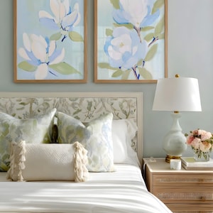 May include: Two framed floral art prints with light blue and white flowers hang above a bed with decorative pillows. A white lamp and a nightstand with a drawer are next to the bed. The room has a light blue wall.