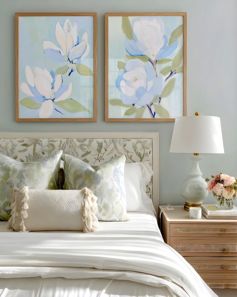 May include: Two framed floral art prints with light blue and white flowers hang above a bed with decorative pillows. A white lamp and a nightstand with a drawer are next to the bed. The room has a light blue wall.