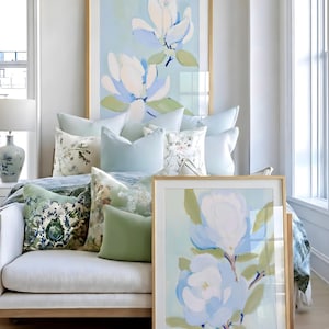 May include: A light-filled room featuring a white sofa adorned with decorative pillows in shades of green and blue. Two framed floral paintings with gold frames lean against the sofa and wall. The artwork depicts large white and blue flowers.