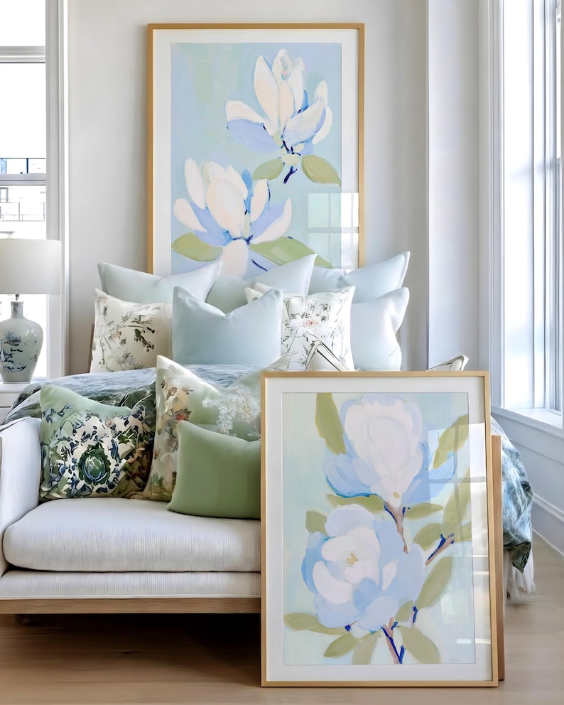 May include: A light-filled room featuring a white sofa adorned with decorative pillows in shades of green and blue. Two framed floral paintings with gold frames lean against the sofa and wall. The artwork depicts large white and blue flowers.