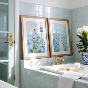 May include: Two framed watercolor paintings of a lighthouse and blue flowers rest on a tiled ledge above a bathtub. The bathroom features a light blue color scheme, with gold fixtures and a white towel.