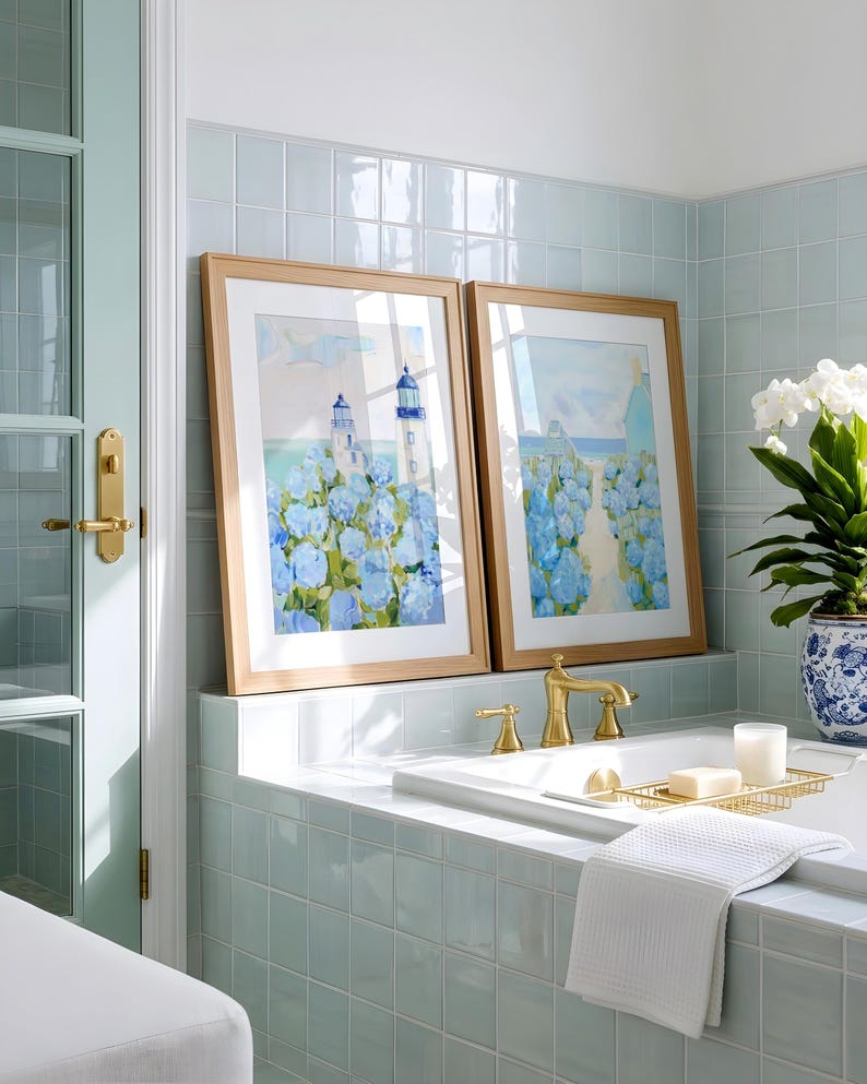 May include: Two framed watercolor paintings of a lighthouse and blue flowers rest on a tiled ledge above a bathtub. The bathroom features a light blue color scheme, with gold fixtures and a white towel.