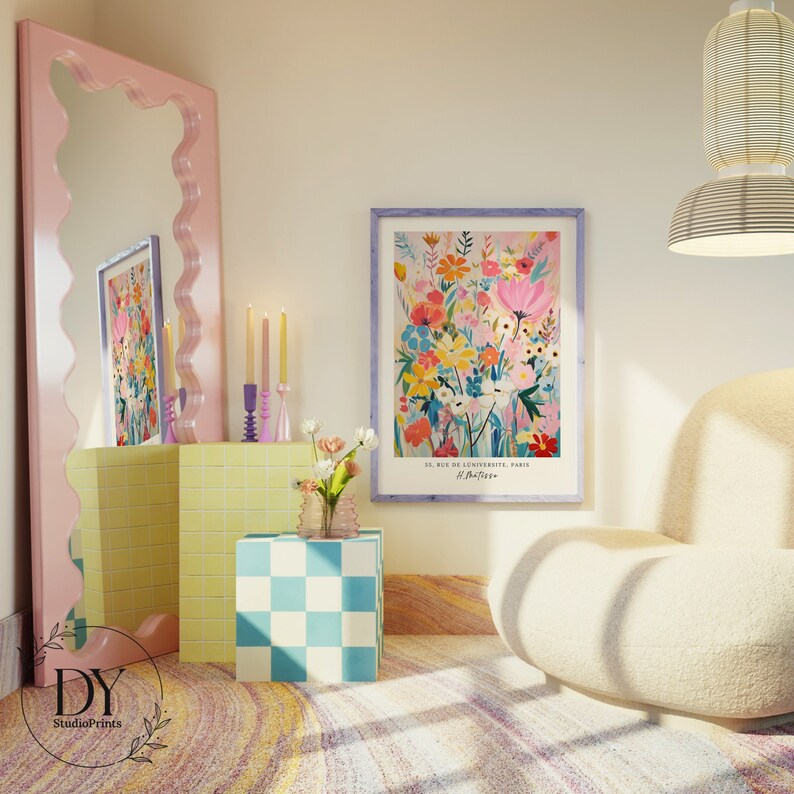 May include: A colorful floral print with a light pink background. The print is framed in a light purple frame and has the text "51, rue de l'Universit&eacute;, Paris" and "#Parisian" at the bottom. The print is hanging on a white wall in a room with a pink mirror, yellow and white tiled shelves, and a white armchair.