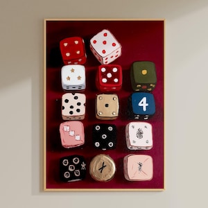Op de afbeelding: Een ingelijst kunstwerk met een verzameling van twaalf geschilderde dobbelstenen tegen een dieprode achtergrond. De dobbelstenen zijn in verschillende kleuren, waaronder rood, wit, zwart en goud, met verschillende punt- en symboolarrangementen. Het kunstwerk is lichtbruin omlijst.