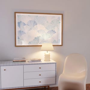 May include: A white dresser with three drawers and a light wood top. The dresser has a white lamp with a cream shade on the top. There are two books on the dresser, one titled "Kinfolk" and the other titled "Nordic Retreat". A white chair is to the right of the dresser. A framed print with a blue and white pattern hangs on the wall above the dresser.