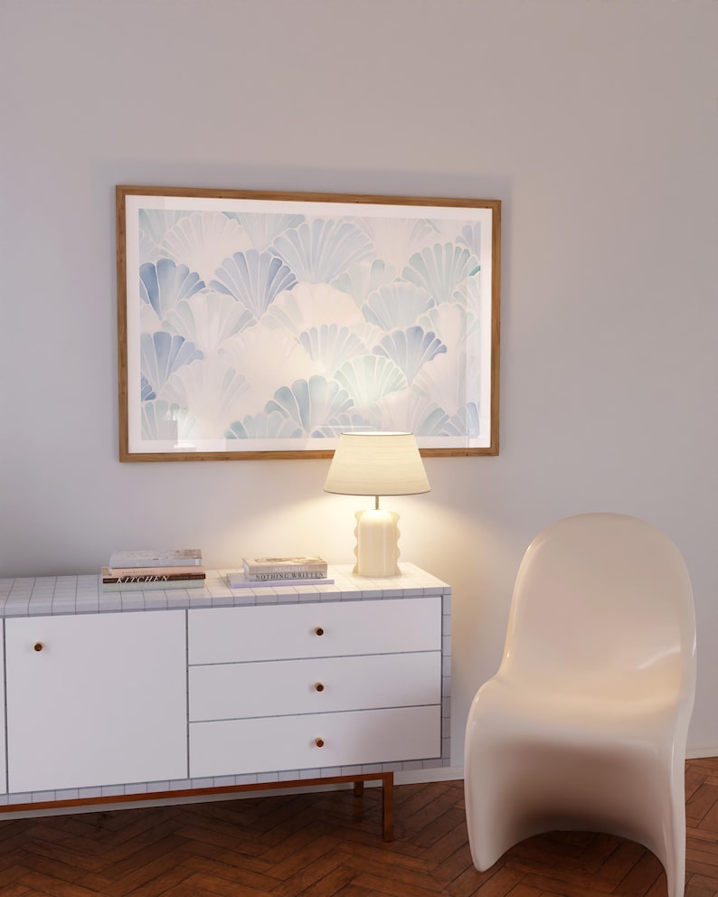 May include: A white dresser with three drawers and a light wood top. The dresser has a white lamp with a cream shade on the top. There are two books on the dresser, one titled "Kinfolk" and the other titled "Nordic Retreat". A white chair is to the right of the dresser. A framed print with a blue and white pattern hangs on the wall above the dresser.