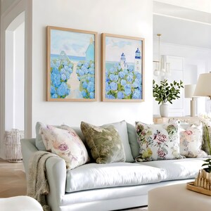 May include: Two framed landscape paintings with blue hydrangeas and a coastal scene hang above a light blue sofa. Decorative pillows with floral and velvet patterns adorn the sofa, along with a neutral-toned throw blanket.