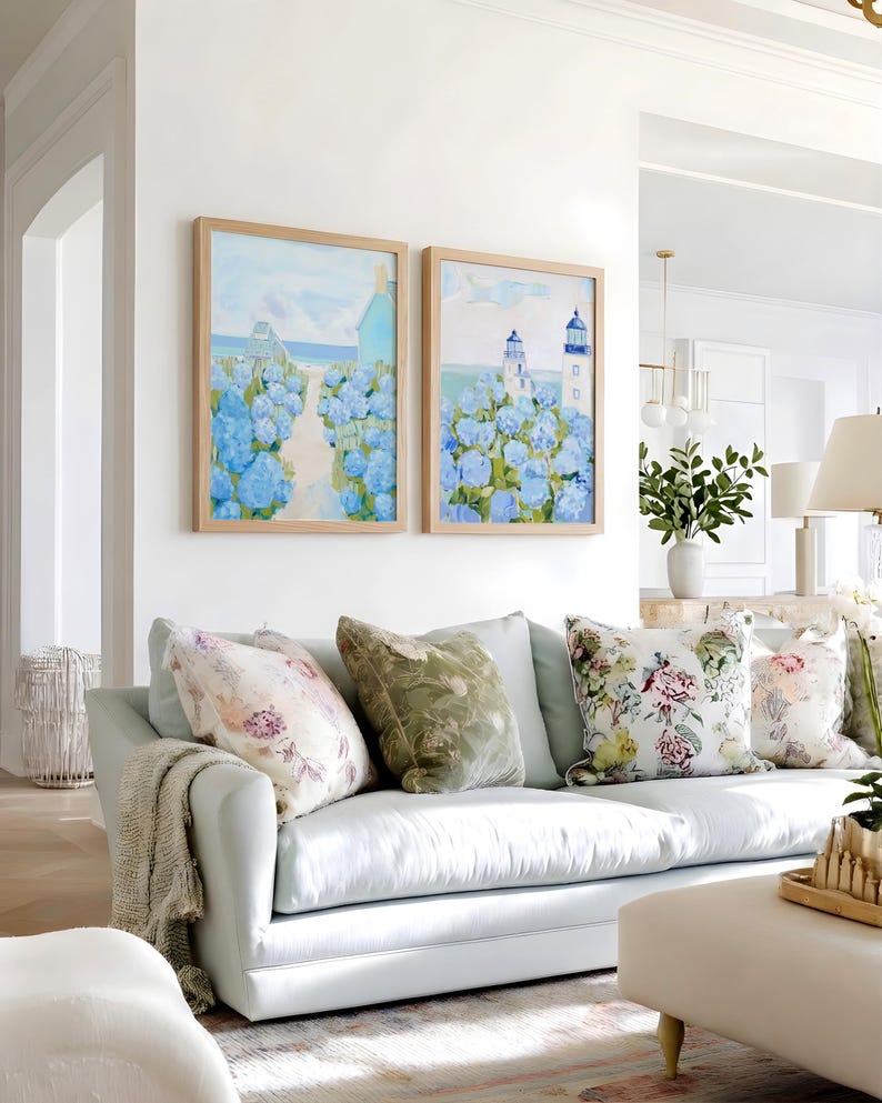 May include: Two framed landscape paintings with blue hydrangeas and a coastal scene hang above a light blue sofa. Decorative pillows with floral and velvet patterns adorn the sofa, along with a neutral-toned throw blanket.