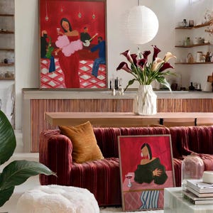 May include: A living room scene featuring a large red velvet sofa with an orange pillow, a framed painting, and a fluffy white ottoman. A vase of calla lilies sits on a wooden table. A large painting hangs on the wall.