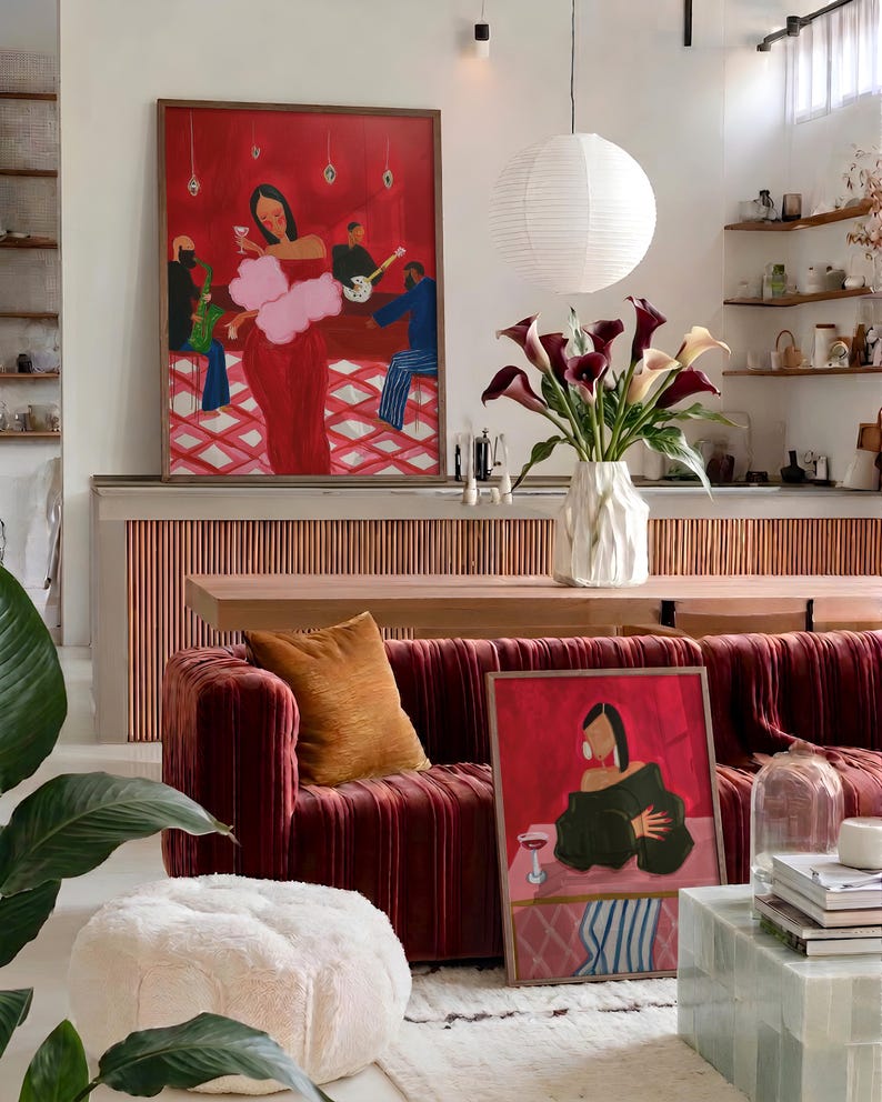 May include: A living room scene featuring a large red velvet sofa with an orange pillow, a framed painting, and a fluffy white ottoman. A vase of calla lilies sits on a wooden table. A large painting hangs on the wall.