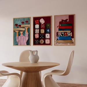 Op de afbeelding: Drie ingelijste kunstdrukken hangen boven een houten tafel en stoelen. De prints tonen een ober met wijnglazen, kleurrijke dobbelstenen en een stapel boeken. Een witte kan staat op de tafel.