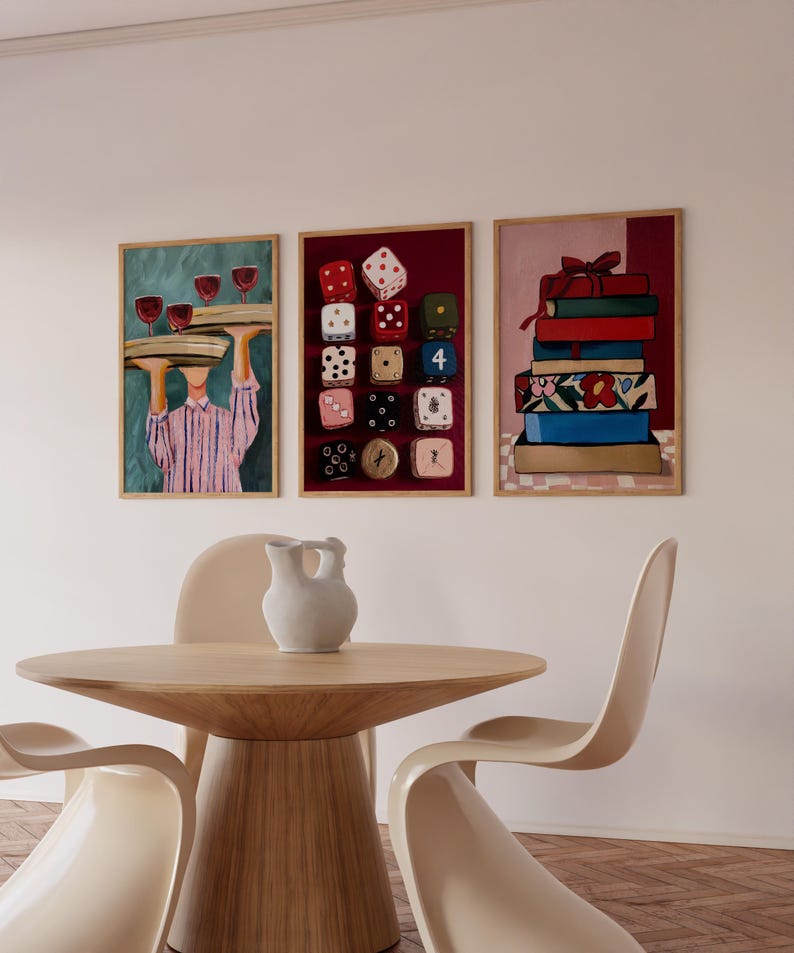 Op de afbeelding: Drie ingelijste kunstdrukken hangen boven een houten tafel en stoelen. De prints tonen een ober met wijnglazen, kleurrijke dobbelstenen en een stapel boeken. Een witte kan staat op de tafel.