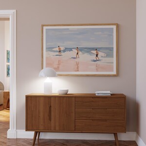 May include: A light wood dresser with three drawers and two doors. The dresser has a white lamp and a white bowl on top. A framed painting of three surfers on a beach hangs on the wall above the dresser.