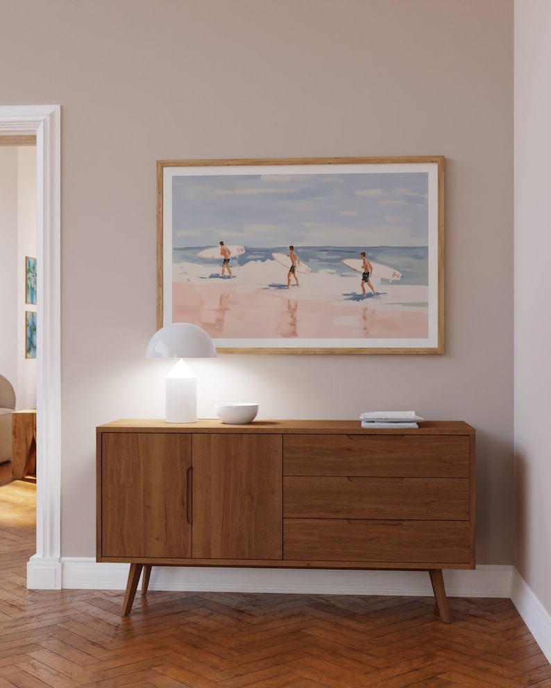May include: A light wood dresser with three drawers and two doors. The dresser has a white lamp and a white bowl on top. A framed painting of three surfers on a beach hangs on the wall above the dresser.