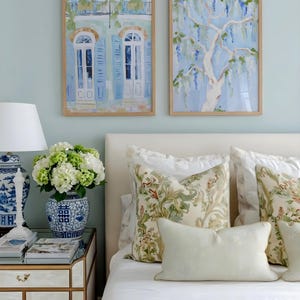Puede incluir: Dos pinturas de acuarela enmarcadas cuelgan sobre una cama con almohadas florales. Una pintura representa un edificio con contraventanas azules, la otra un &aacute;rbol con flores azules en cascada. Un jarr&oacute;n azul y blanco contiene flores blancas y verdes.