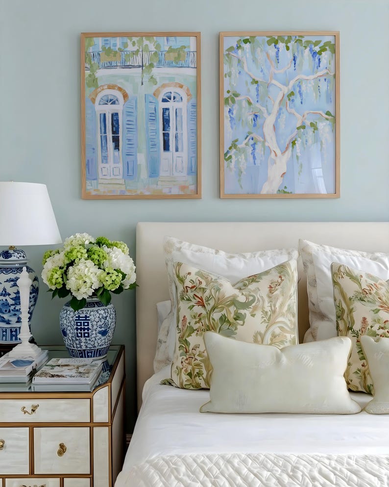 Puede incluir: Dos pinturas de acuarela enmarcadas cuelgan sobre una cama con almohadas florales. Una pintura representa un edificio con contraventanas azules, la otra un &aacute;rbol con flores azules en cascada. Un jarr&oacute;n azul y blanco contiene flores blancas y verdes.