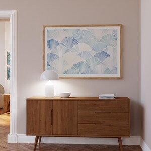 May include: A mid-century modern style wooden sideboard with two doors and three drawers. The sideboard is a light brown wood with a natural finish. A white lamp with a white shade is on top of the sideboard, along with a white bowl. A framed print with a blue and white abstract design is hanging on the wall above the sideboard.