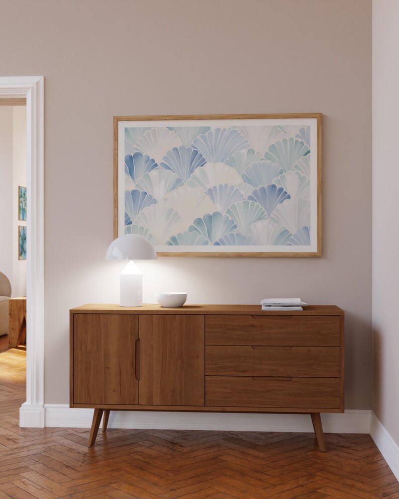 May include: A mid-century modern style wooden sideboard with two doors and three drawers. The sideboard is a light brown wood with a natural finish. A white lamp with a white shade is on top of the sideboard, along with a white bowl. A framed print with a blue and white abstract design is hanging on the wall above the sideboard.