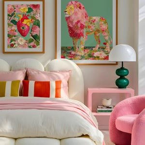 K&ouml;nnte beinhalten: Schlafzimmer-Interieur mit gerahmten Kunstdrucken, einem Bett mit rosa und gestreiften Kissen und einem rosa Nachttisch. Die Kunstwerke umfassen ein Blumenherz und einen L&ouml;wen mit floraler M&auml;hne. Eine gr&uuml;ne Lampe und ein rosa Stuhl vervollst&auml;ndigen die Einrichtung.