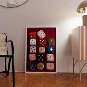 Op de afbeelding: Een ingelijst kunstwerk met twaalf kleurrijke dobbelstenen tegen een dieprode achtergrond. De dobbelstenen hebben verschillende kleuren, waaronder rood, wit, groen en roze, met verschillende punt- en symboolarrangementen. Het kunstwerk wordt getoond in een kamer met een houten vloer.