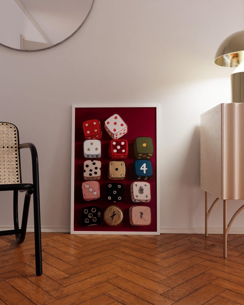 Op de afbeelding: Een ingelijst kunstwerk met twaalf kleurrijke dobbelstenen tegen een dieprode achtergrond. De dobbelstenen hebben verschillende kleuren, waaronder rood, wit, groen en roze, met verschillende punt- en symboolarrangementen. Het kunstwerk wordt getoond in een kamer met een houten vloer.
