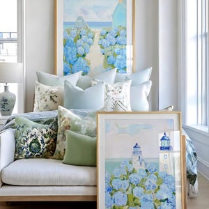 May include: A cozy living space featuring framed art with coastal scenes, including a lighthouse and a path to the beach. The room is decorated with light blue and green throw pillows, a white sofa, and a wooden floor.