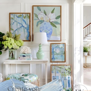 May include: A gallery wall featuring framed art prints with botanical and landscape themes. The artwork includes a large floral painting, a tree scene, and a house with a blue and white color scheme. A white console table holds a lamp and decorative items.