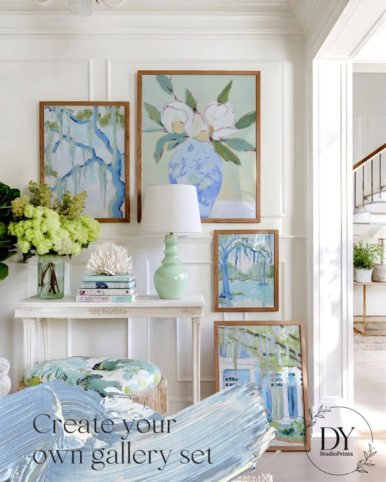 May include: A gallery wall featuring framed art prints with botanical and landscape themes. The artwork includes a large floral painting, a tree scene, and a house with a blue and white color scheme. A white console table holds a lamp and decorative items.