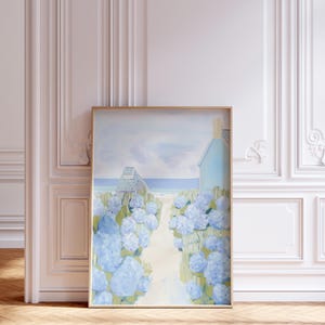 May include: Framed art print depicting a coastal scene. A path leads to a beach and a light blue house, lined with blue hydrangea bushes. The artwork is in a gold frame, set against a white wall and wooden floor.