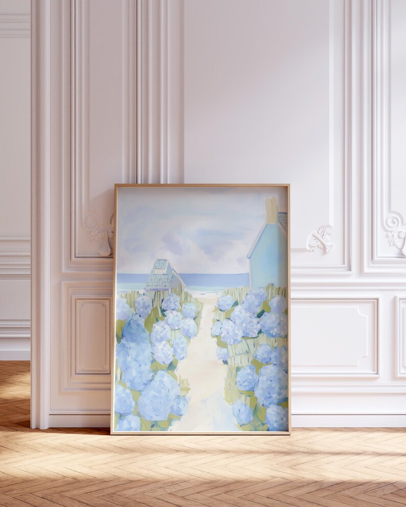 May include: Framed art print depicting a coastal scene. A path leads to a beach and a light blue house, lined with blue hydrangea bushes. The artwork is in a gold frame, set against a white wall and wooden floor.