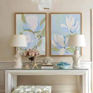 May include: A living room scene featuring two framed floral paintings with light blue and white flowers. A white console table holds two white lamps, a vase of flowers, and decorative items. A patterned ottoman sits below the table.
