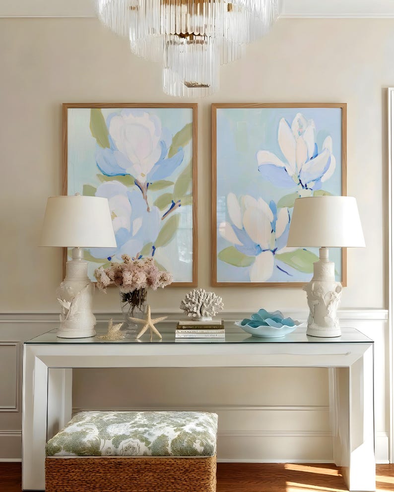 May include: A living room scene featuring two framed floral paintings with light blue and white flowers. A white console table holds two white lamps, a vase of flowers, and decorative items. A patterned ottoman sits below the table.