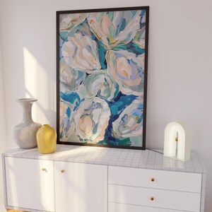 K&ouml;nnte beinhalten: Eine wei&szlig;e Kommode mit drei Schubladen und goldenen Kn&ouml;pfen. Die Kommode ist mit einem schwarzen gerahmten abstrakten Gem&auml;lde von blauen und wei&szlig;en Blumen verziert. Eine graue Vase und eine gelbe Vase stehen auf der linken Seite der Kommode. Eine wei&szlig;e, bogenf&ouml;rmige Lampe steht auf der rechten Seite der Kommode.