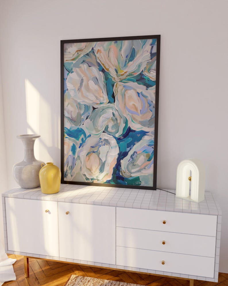 K&ouml;nnte beinhalten: Eine wei&szlig;e Kommode mit drei Schubladen und goldenen Kn&ouml;pfen. Die Kommode ist mit einem schwarzen gerahmten abstrakten Gem&auml;lde von blauen und wei&szlig;en Blumen verziert. Eine graue Vase und eine gelbe Vase stehen auf der linken Seite der Kommode. Eine wei&szlig;e, bogenf&ouml;rmige Lampe steht auf der rechten Seite der Kommode.