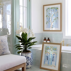 Puede incluir: Un interior de ba&ntilde;o con obras de arte en acuarela enmarcadas, una orqu&iacute;dea en maceta y un banco con un coj&iacute;n estampado. Las obras de arte representan un &aacute;rbol y un edificio con contraventanas azules. La habitaci&oacute;n tiene una combinaci&oacute;n de colores azul claro y blanco.