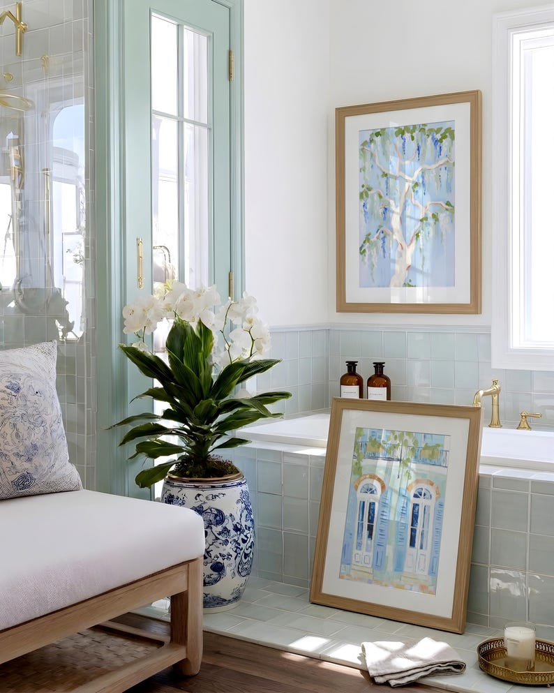 Puede incluir: Un interior de ba&ntilde;o con obras de arte en acuarela enmarcadas, una orqu&iacute;dea en maceta y un banco con un coj&iacute;n estampado. Las obras de arte representan un &aacute;rbol y un edificio con contraventanas azules. La habitaci&oacute;n tiene una combinaci&oacute;n de colores azul claro y blanco.