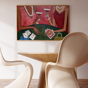 May include: An abstract painting depicts two figures in pink and red dresses, holding cocktails, with playing cards on a green table. The artwork is framed and hangs above a wooden table and two cream-colored chairs.