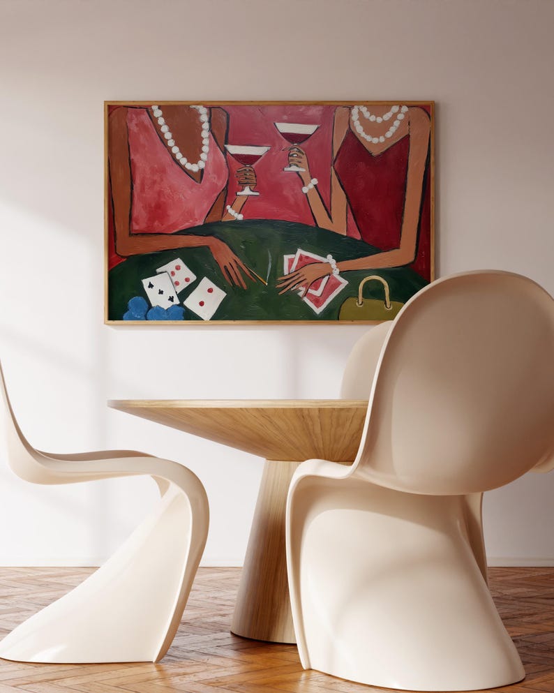 May include: An abstract painting depicts two figures in pink and red dresses, holding cocktails, with playing cards on a green table. The artwork is framed and hangs above a wooden table and two cream-colored chairs.