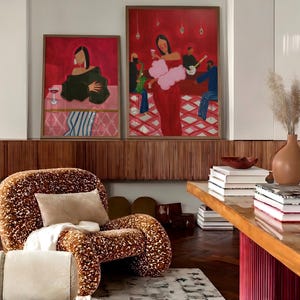 May include: Two framed paintings with a red background and figures in a bar setting hang above a patterned chair. The chair has a brown and cream pattern. A wooden table with books and a vase is in the foreground.
