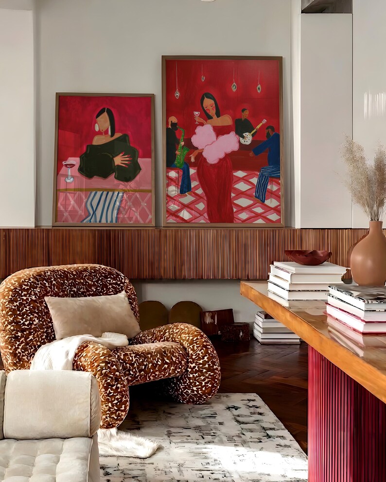May include: Two framed paintings with a red background and figures in a bar setting hang above a patterned chair. The chair has a brown and cream pattern. A wooden table with books and a vase is in the foreground.