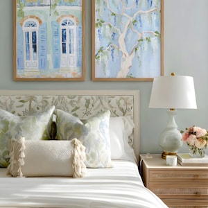 Puede incluir: Escena de dormitorio con dos pinturas de acuarela enmarcadas, una de un edificio con contraventanas azules y otra de un &aacute;rbol con flores azules. La cama tiene almohadas estampadas y un edred&oacute;n blanco. Una l&aacute;mpara y una mesita de noche con flores completan la escena.