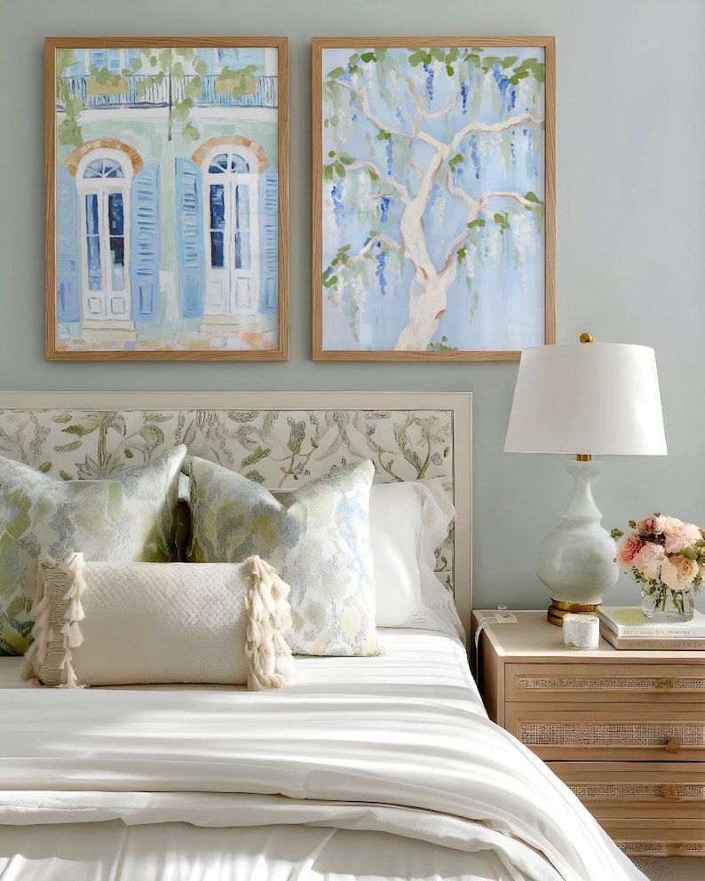 Puede incluir: Escena de dormitorio con dos pinturas de acuarela enmarcadas, una de un edificio con contraventanas azules y otra de un &aacute;rbol con flores azules. La cama tiene almohadas estampadas y un edred&oacute;n blanco. Una l&aacute;mpara y una mesita de noche con flores completan la escena.