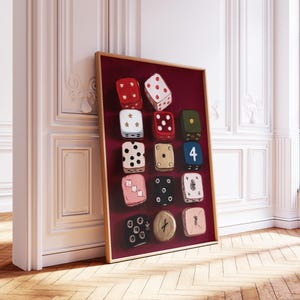 Op de afbeelding: Een ingelijst kunstwerk met een verzameling van twaalf dobbelstenen tegen een dieprode achtergrond. De dobbelstenen zijn geschilderd in verschillende kleuren, waaronder rood, zwart en roze, met witte ogen. Het kunstwerk wordt tentoongesteld in een kamer met witte muren en een houten vloer.