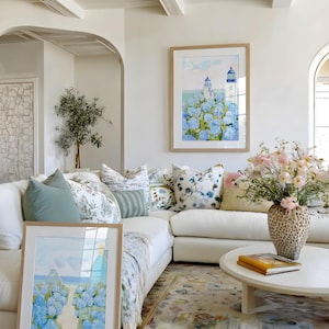 May include: A bright, airy living room with a white sectional sofa, decorative pillows, and a round coffee table. Two framed watercolor paintings of lighthouses and blue flowers are displayed. A vase of pink flowers sits on the table, and a patterned rug covers the floor.