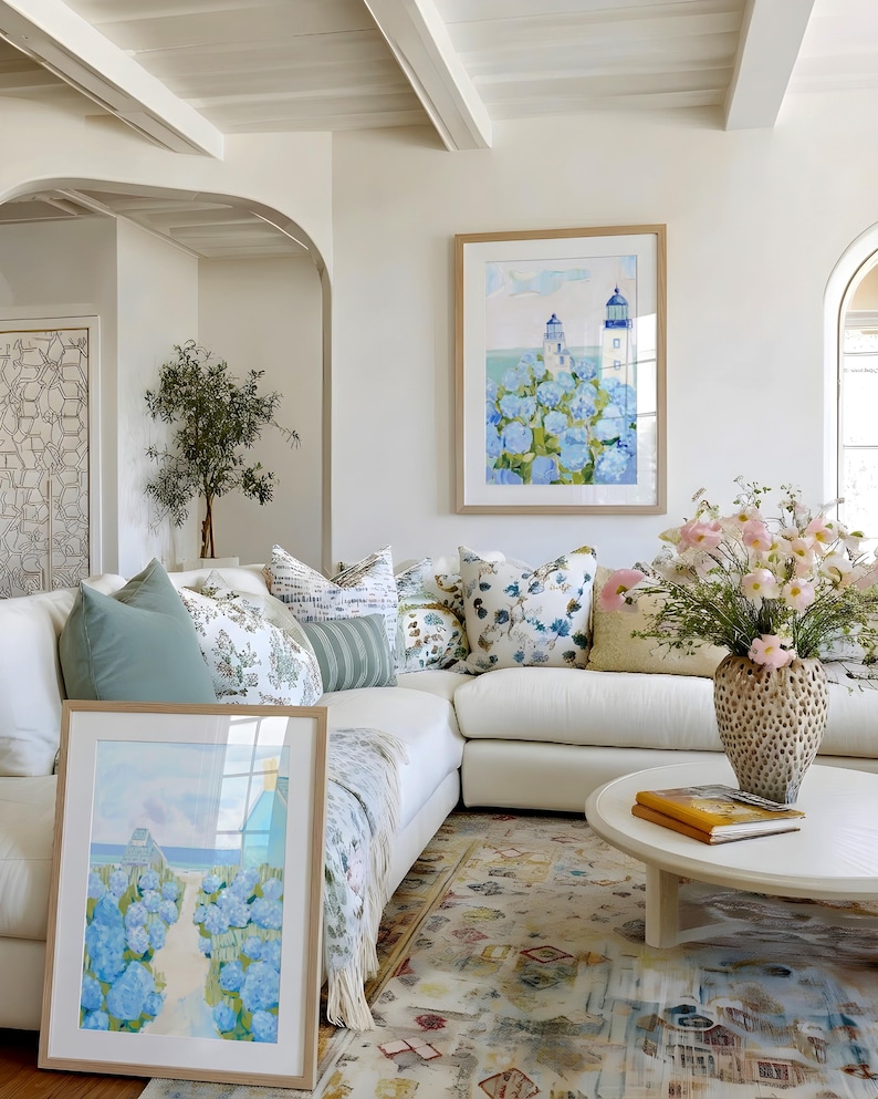 May include: A bright, airy living room with a white sectional sofa, decorative pillows, and a round coffee table. Two framed watercolor paintings of lighthouses and blue flowers are displayed. A vase of pink flowers sits on the table, and a patterned rug covers the floor.