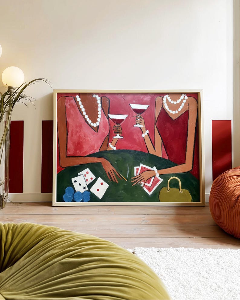 May include: An abstract painting depicts two figures in red dresses, each holding a martini glass. They are seated at a green table with playing cards, poker chips, and a purse. The artwork is framed in a light wood.