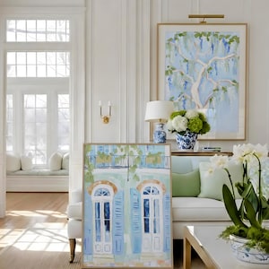Puede incluir: Escena interior con dos pinturas enmarcadas, un sof&aacute; blanco con cojines verdes y una mesa de centro blanca con patas doradas. Una pintura representa un &aacute;rbol con follaje azul y verde, la otra una fachada de edificio con contraventanas azules.