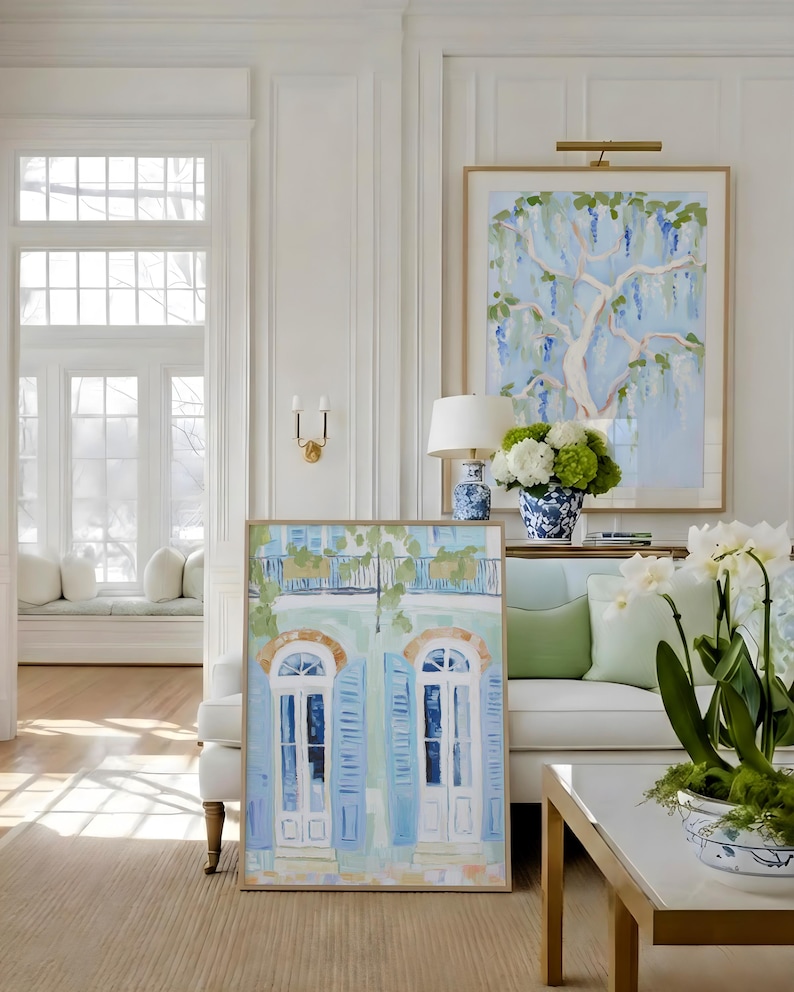 Puede incluir: Escena interior con dos pinturas enmarcadas, un sof&aacute; blanco con cojines verdes y una mesa de centro blanca con patas doradas. Una pintura representa un &aacute;rbol con follaje azul y verde, la otra una fachada de edificio con contraventanas azules.