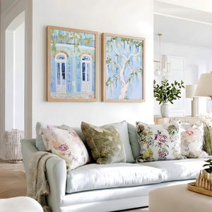Puede incluir: Un sof&aacute; azul claro con cojines florales y estampados y una manta beige. Dos pinturas enmarcadas cuelgan sobre el sof&aacute;, una que representa un edificio con contraventanas azules y la otra un &aacute;rbol con flores colgantes. La habitaci&oacute;n tiene un ambiente luminoso y aireado.